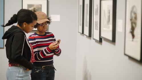 Two young women stand in the galleries, looking at something unseen one of them is holding in her hands