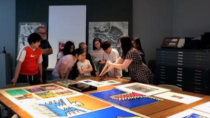 Large prints lay on a table, while a curator holds a work to show a group of students